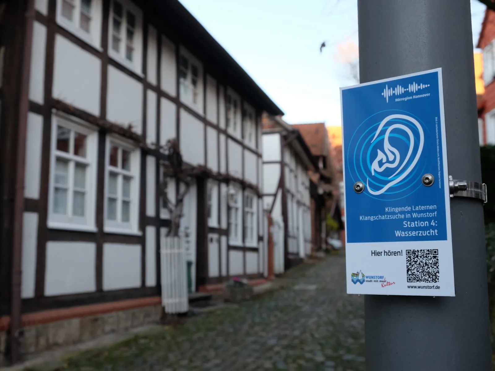 Ein blaues Schild mit einem gezeichneten Ohr und einem QR-Code an einem Laternenpfahl